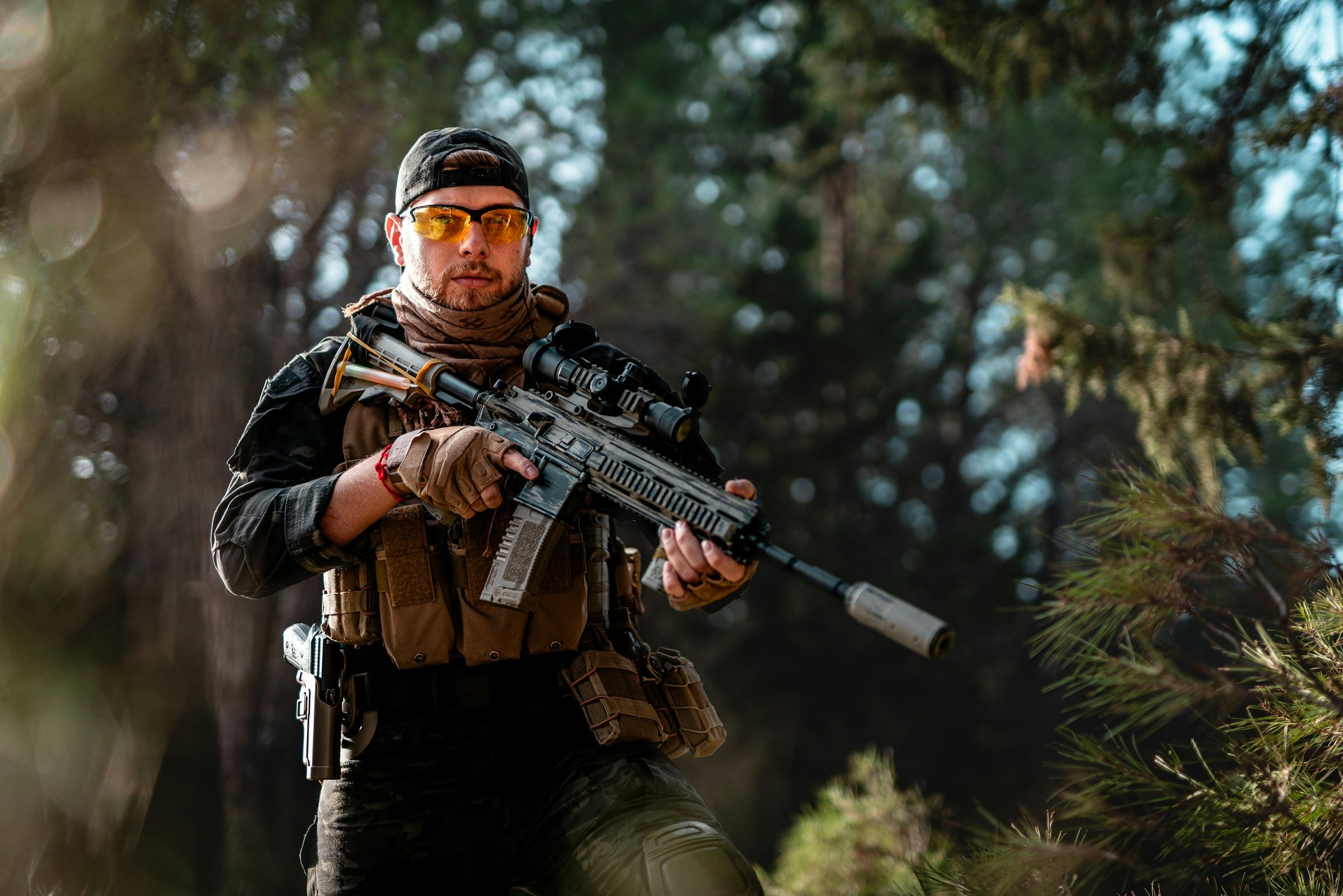 An adult male surrounded by the forest with his tactical gear on. 